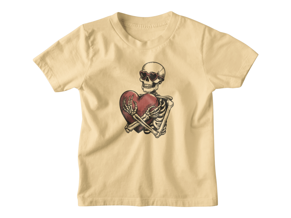 A light yellow short-sleeve kids t-shirt with a Halloween graphic of a skeleton wearing sunglasses and holding a broken red heart, displayed on a clean white background.