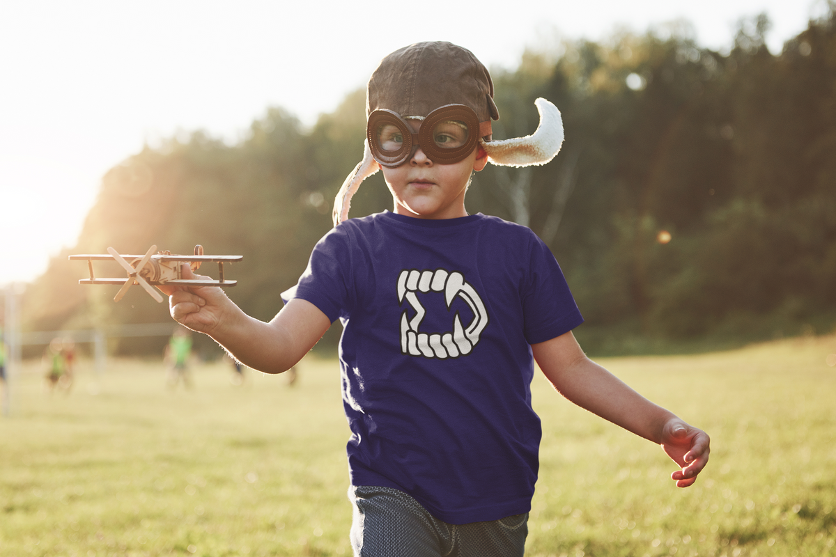 A young boy wearing aviator goggles, running in a grassy park wearing a purple shirt with a graphic of Halloween plastic vampire teeth.