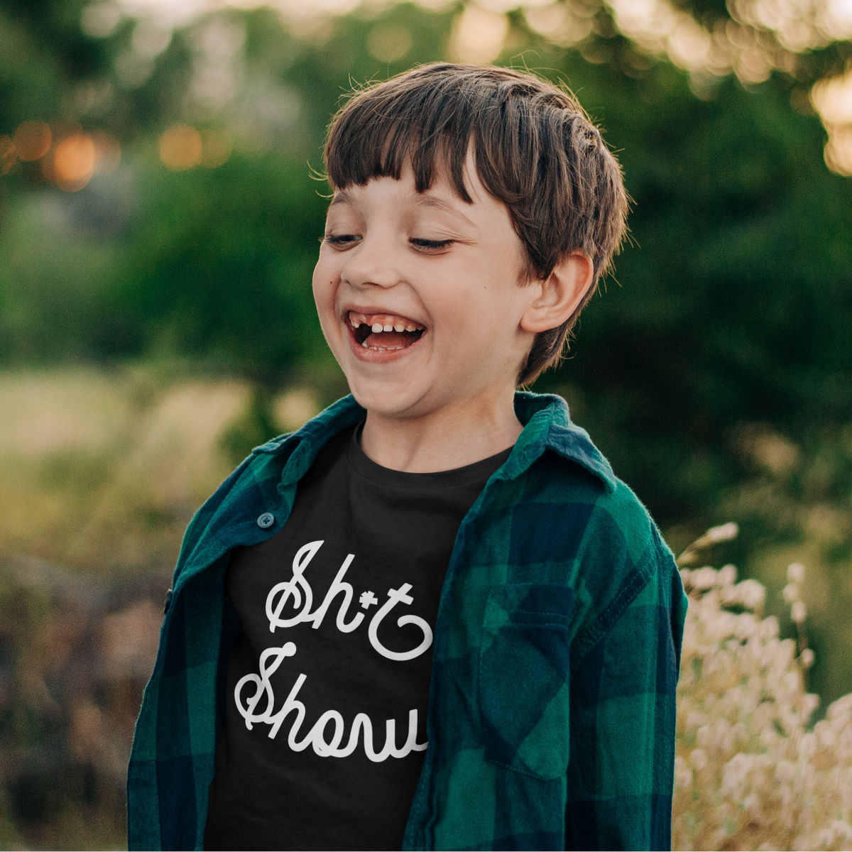 A young boy laughing outdoors, wearing a black graphic t-shirt with white cursive text that reads 'Sh*t Show', layered under a green and black plaid flannel shirt.