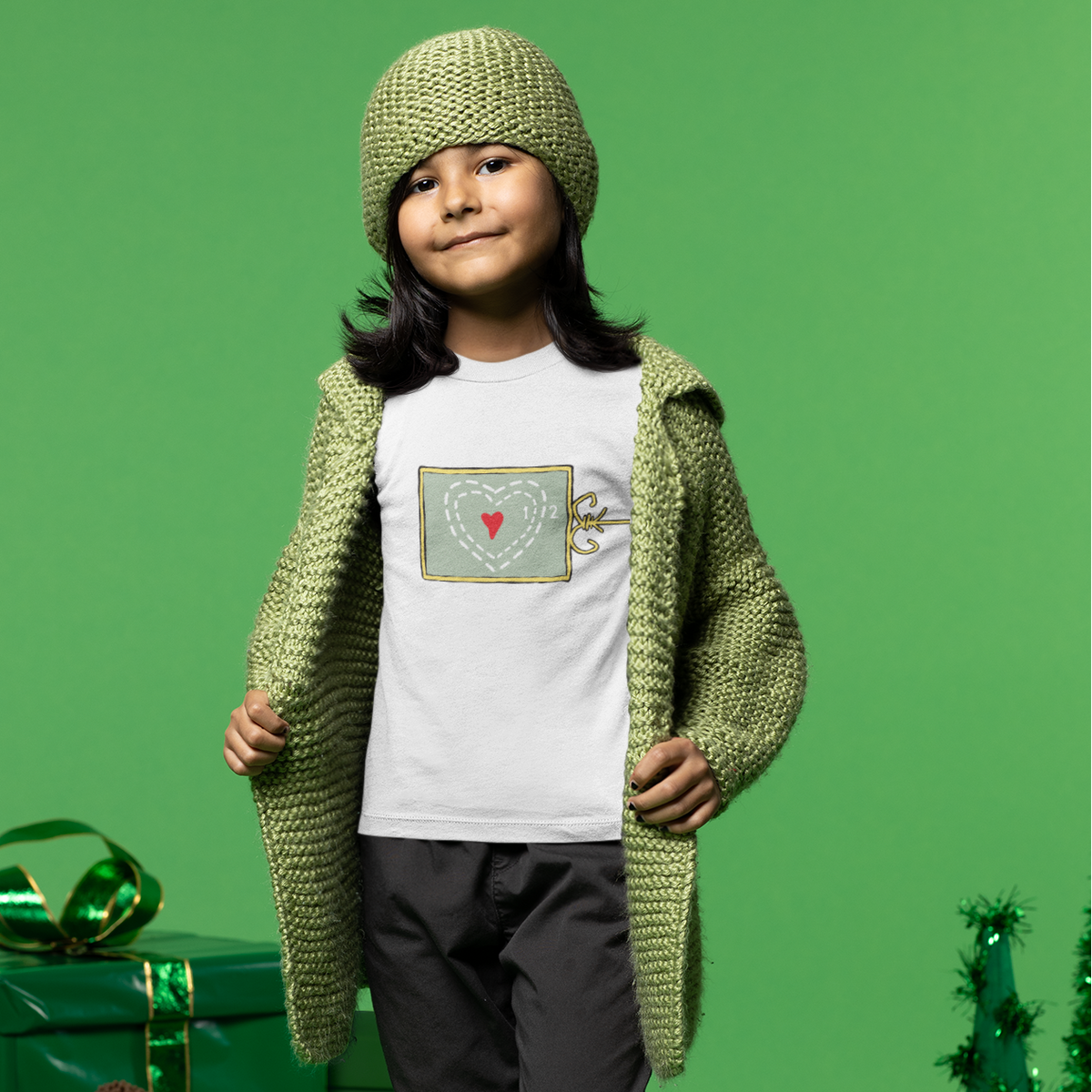 A child model wearing a white graphic t-shirt, a green knit cardigan, a matching green knit beanie, dark gray pants, and black boots, standing against a solid green studio background with Christmas-themed props.