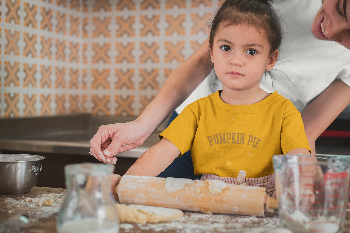 A young girl wearing a yellow toddler t-shirt with a 'Pumpkin Pie' Thanksgiving theme sitting at a wooden table covered in flour.