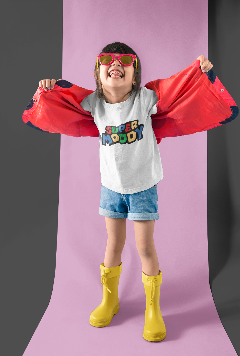 A kid modeling a white graphic t-shirt with 'SUPER MOODY' in Mario Brothers style. Paired with denim shorts, bright yellow rain boots, and pink sunglasses.