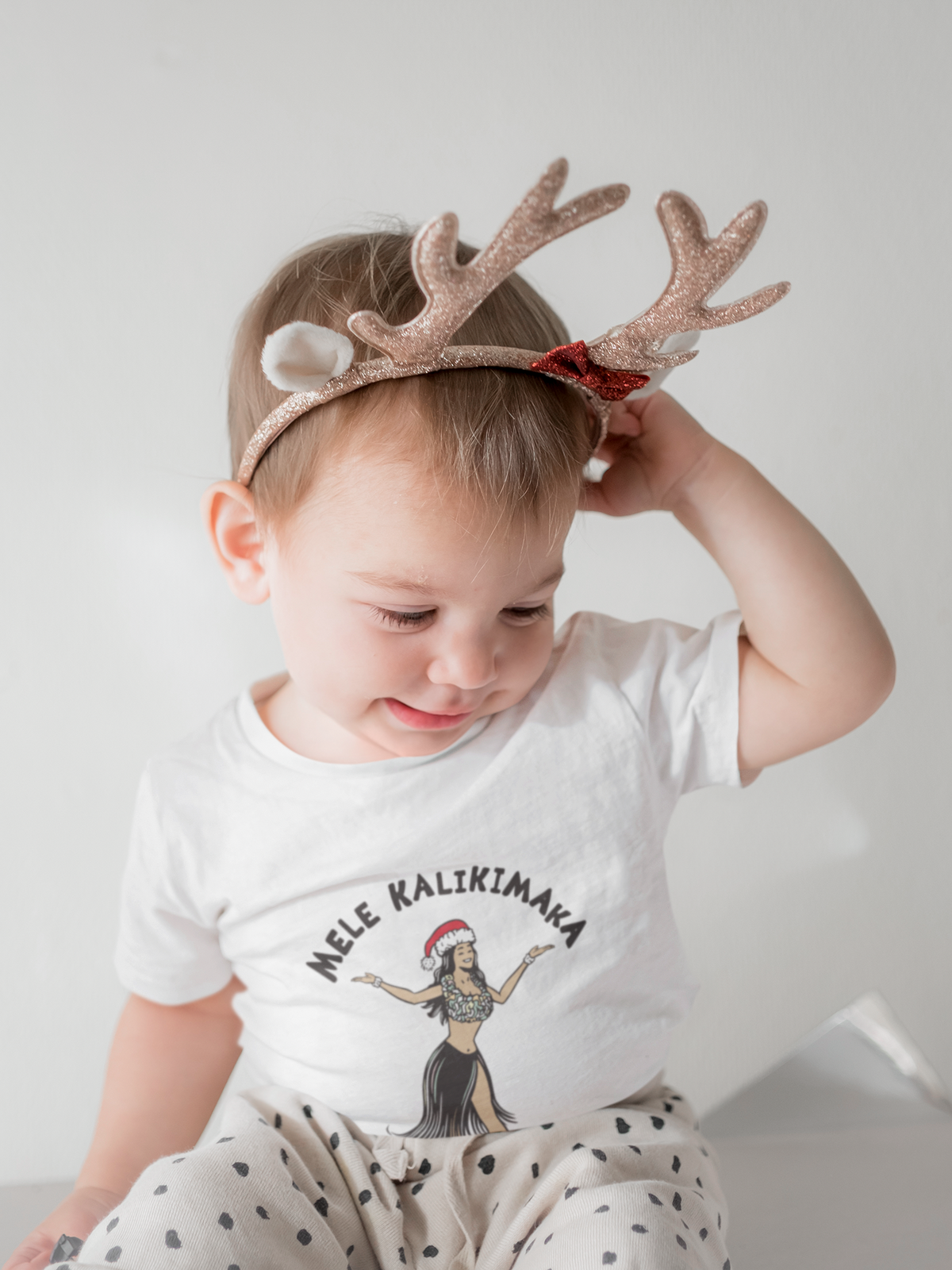 A toddler wearing a Christmas reindeer antler headband with a white t-shirt that reads 'MELE KALIKIMAKA' which means Hawaiian Merry Christmas.