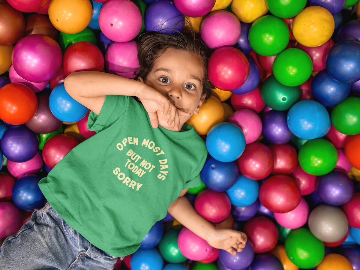 Young child in a ball pit with a green shirt that has a caption of 'Open Most Days But Not Today Sorry'.